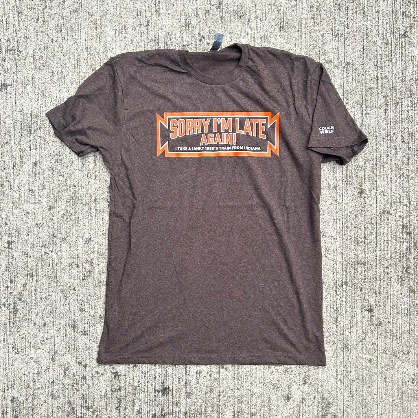 A dark brown heather short-sleeve t-shirt laid flat on a textured gray concrete surface. The shirt features a graphic on the chest with orange and white text reading "SORRY I'M LATE AGAIN' and 'I TAKE A JANKY 1980's TRAIN FROM INDIANA", plus a small white couch wolf logo on the left sleeve.