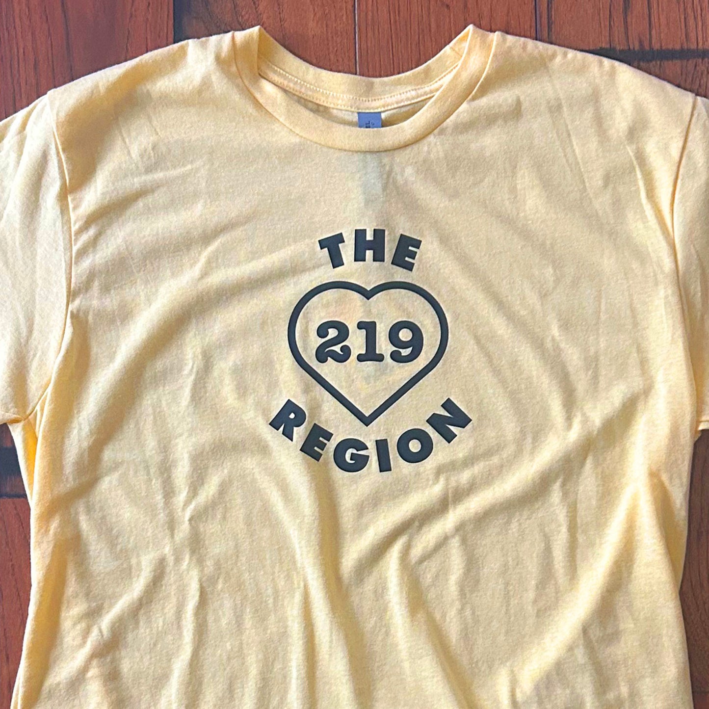 The Region 219 shirt northwest indiana t-shirt says the region 219 inside of a heart yellow short sleeve shirt on wood floor background