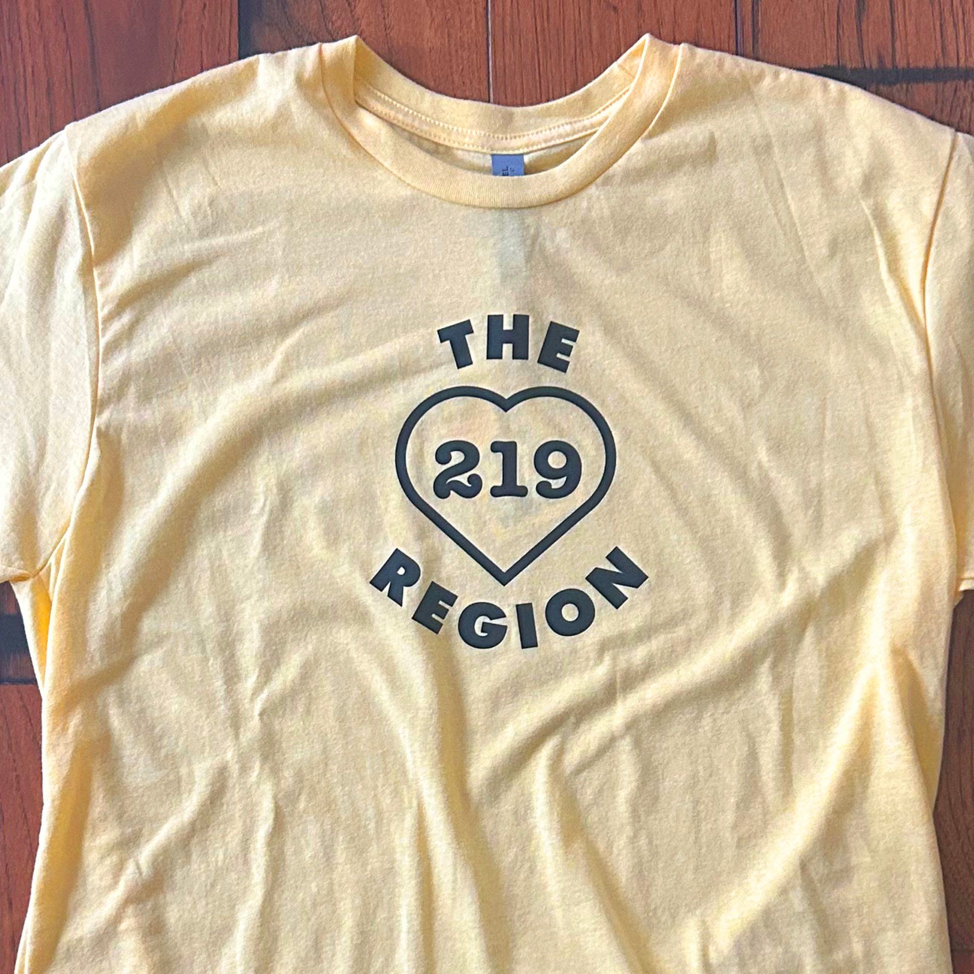 The Region 219 shirt northwest indiana t-shirt says the region 219 inside of a heart yellow short sleeve shirt on wood floor background
