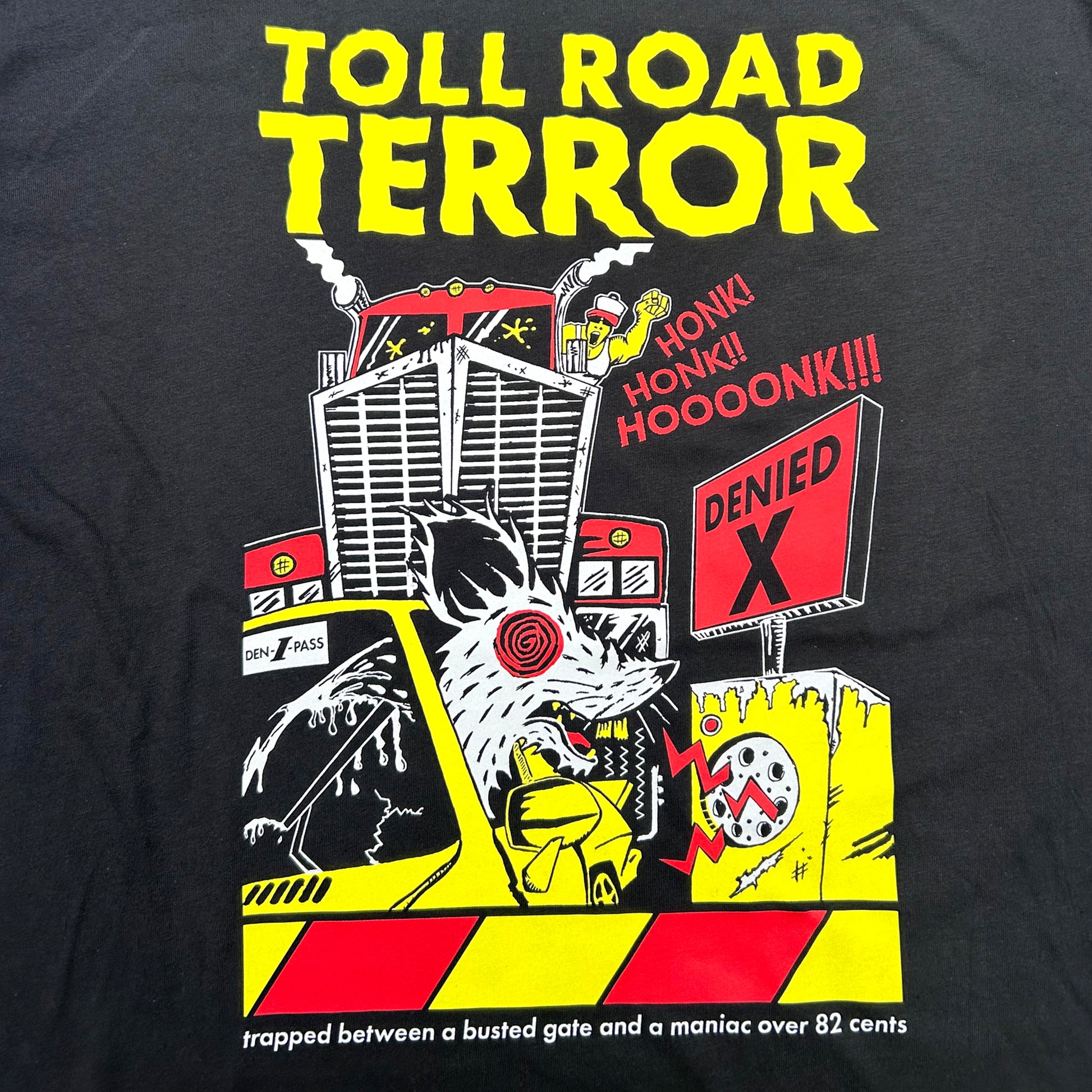 Close-up of a black graphic T-shirt featuring a bold yellow and red 'TOLL ROAD TERROR' print with cartoon vehicles, a rat, and warning-style artwork.