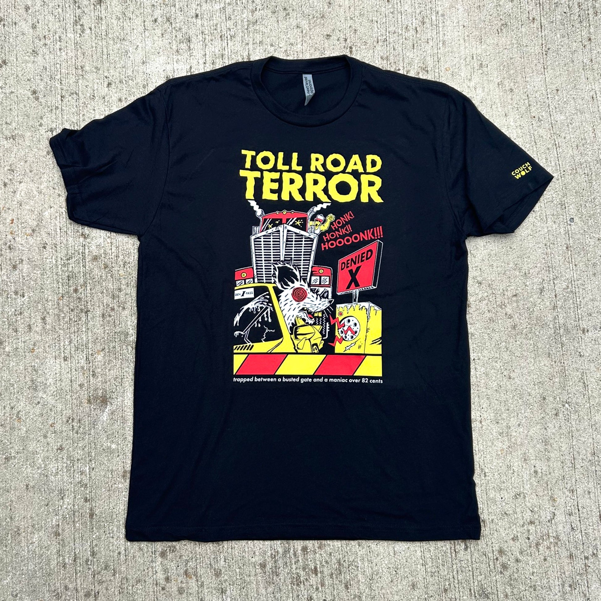 A black short-sleeve graphic t-shirt laid flat on a textured gray concrete surface. The graphic features the words 'TOLL ROAD TERROR' in bold yellow letters above a collage artwork of rar yelling into speaker as a semi truck honks behind him.