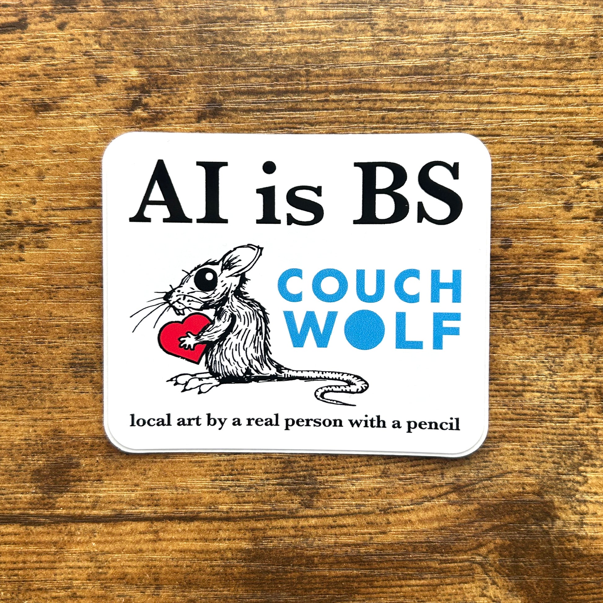 A square white sticker with rounded corners lies on a rustic brown wooden surface. The sticker features the text 'AI is BS' in black, an illustration of a black-and-white rat holding a red heart, the words 'COUCH WOLF' in blue, and the phrase 'local art by a real person with a pencil' in black at the bottom.