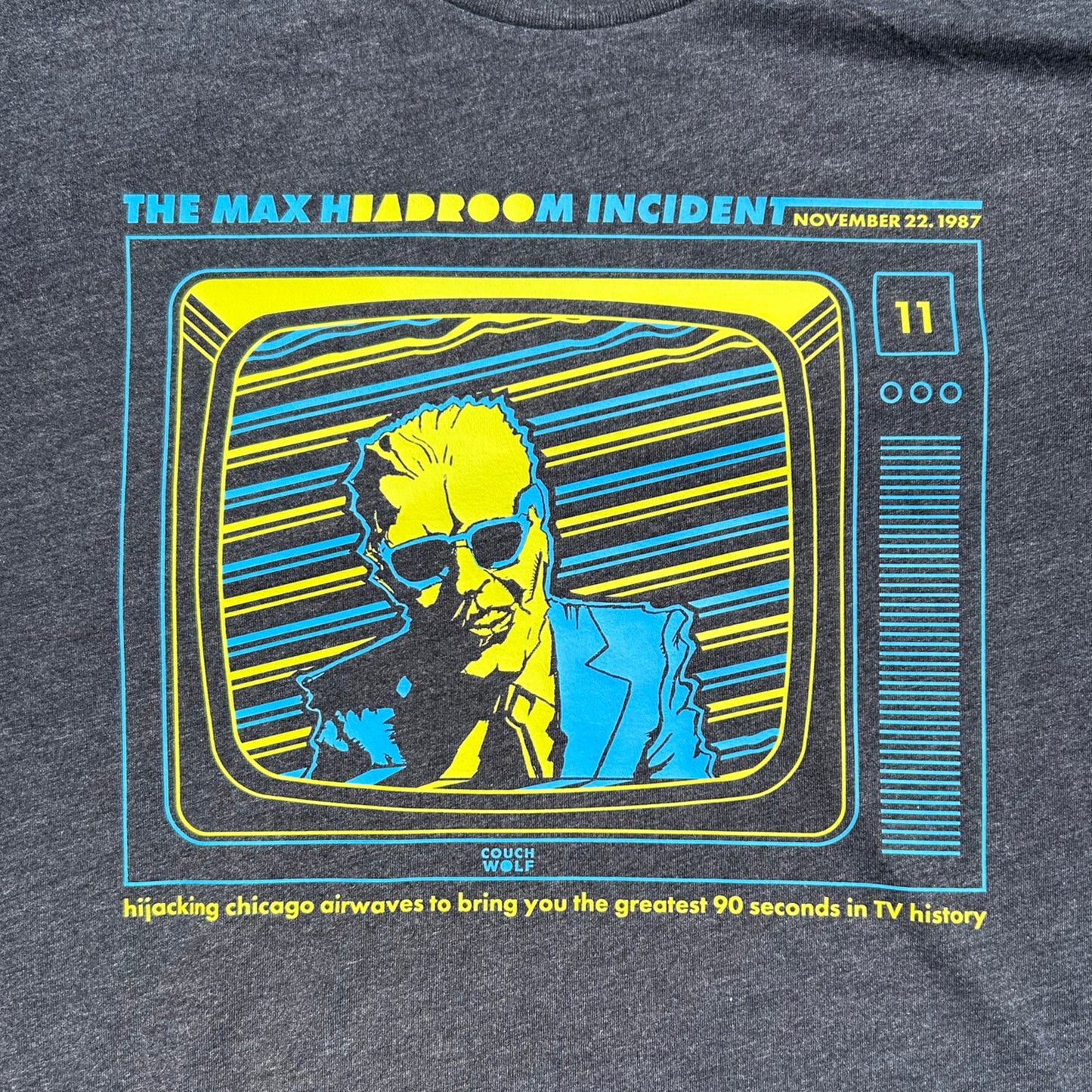 Close-up of a dark heather gray t-shirt featuring a retro graphic print of a man in sunglasses on a stylized television screen, with text reading 'THE MAX INCIDENT' and 'Couch Wolf'.