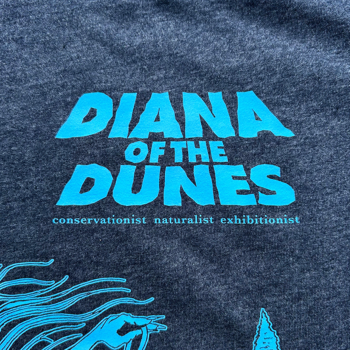 Close-up of a dark heather navy t-shirt featuring a bright blue graphic print that reads 'DIANA OF THE DUNES' with the subtitle 'conservationist naturalist exhibitionist' and partial graphic on bottom