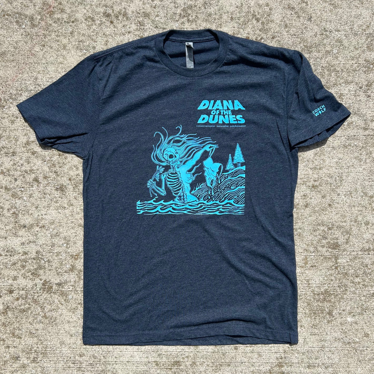 A navy blue heather t-shirt with a bright blue graphic print of 'Diana of the Dunes' on the front, laid flat on a textured gray concrete surface.