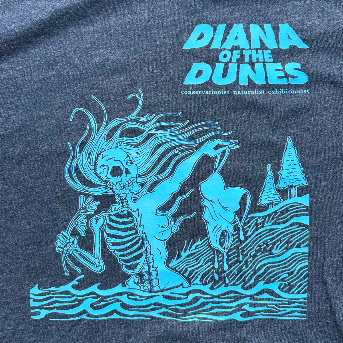 Close-up of a dark heather navy t-shirt featuring a bright blue  graphic print of a skeleton ghost standing in water next to a sand dune with the text that says  'DIANA OF THE DUNES' and smaller text 'conservationist naturalist exhibitionist'.