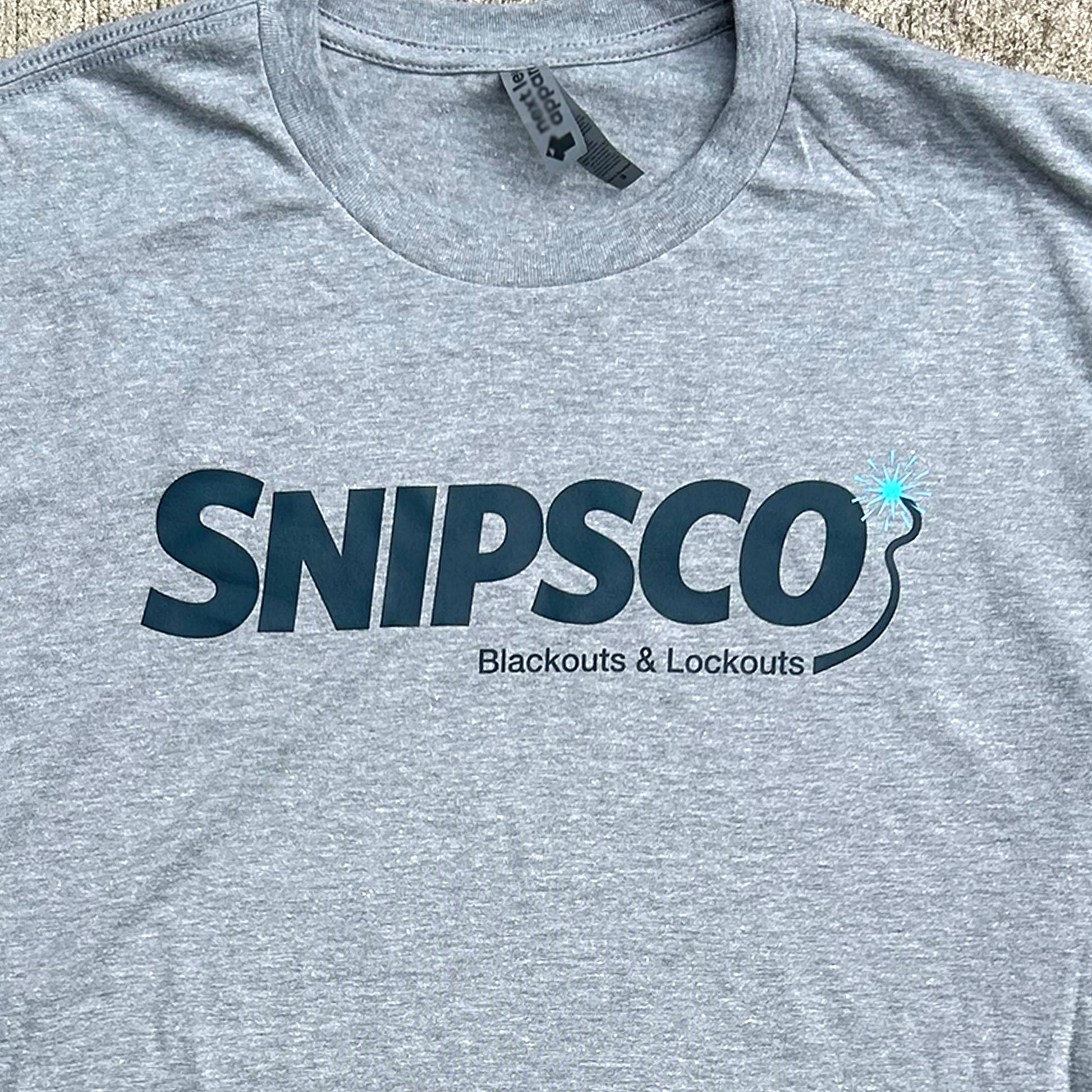 A close-up of a heather gray t-shirt featuring a dark navy blue print that reads 'snips-co Blackouts & Lockouts' with an arcing wire