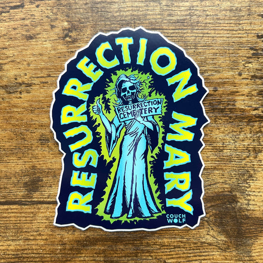 A die-cut sticker featuring a stylized skeleton figure in a blue dress holding a sign that reads 'RESURRECTION MARY' surrounded by large yellow-green text arched over the top and sides. The sticker is photographed on a rustic wooden surface.