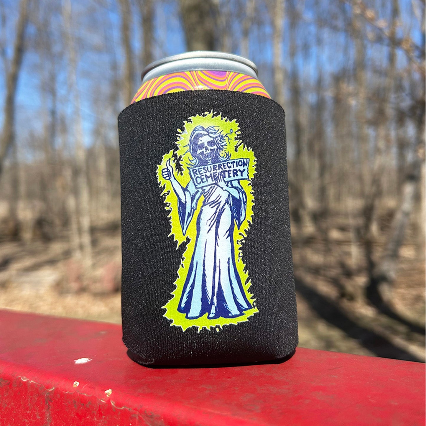 Resurrection Mary Ghost can cooler foam can cooler blue and green color artwork of hitchhiking ghost resurrection Mary holding sign with thumb out that  says "Resurrection Cemetery" on red surface winter woods in background, rainbow can inside can cooler